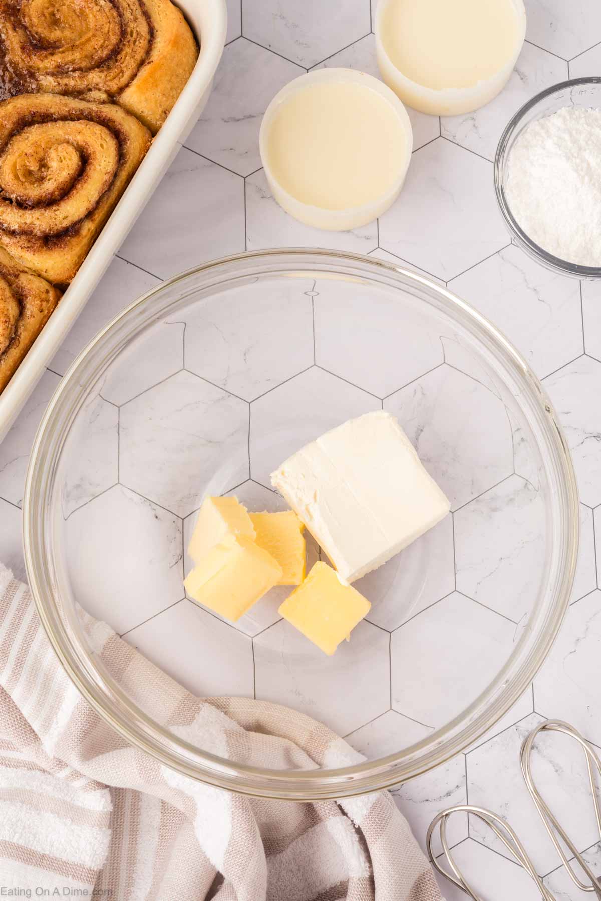 A glass bowl with cream cheese and butter cubes sits on a hexagon-tiled counter, surrounded by a dish of TikTok Cinnamon Rolls Recipe, two small bowls of milk, powdered sugar, a striped towel, and a hand mixer.