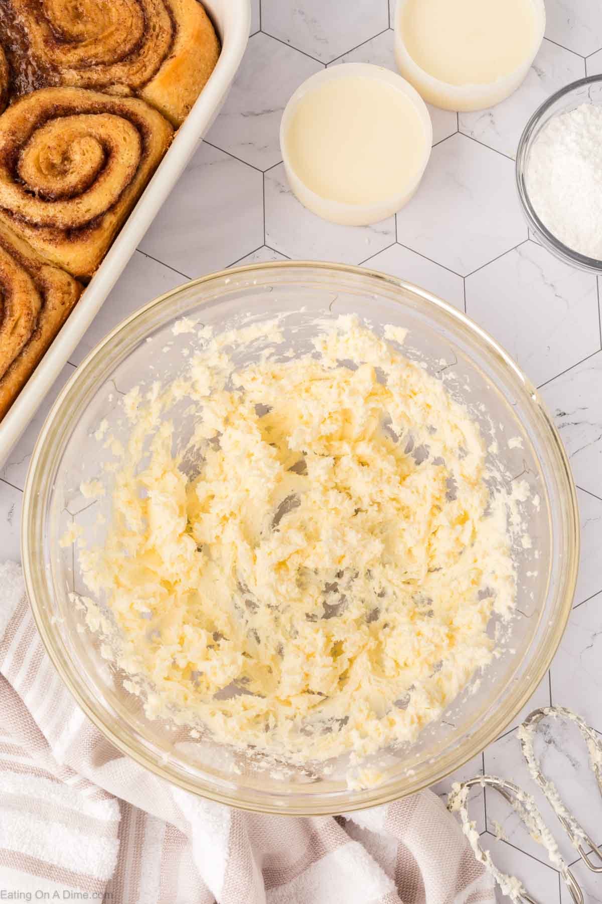 A glass bowl filled with creamed butter and sugar sits on a marble counter, surrounded by a baking dish of TikTok Cinnamon Rolls Recipe, two small bowls of milk, and a bowl of powdered sugar, with a striped cloth and mixer beaters nearby.