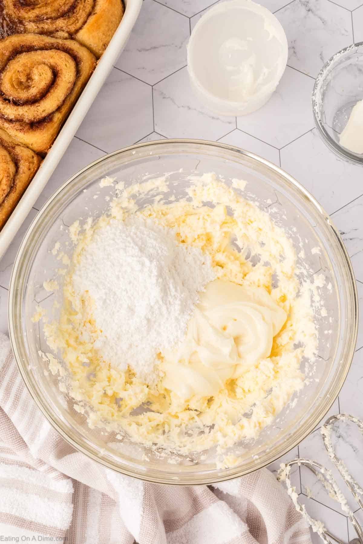 A glass bowl contains softened butter, cream cheese, and powdered sugar, ready to be mixed for a TikTok Cinnamon Rolls Recipe. Nearby are cinnamon rolls in a baking dish, a small bowl of frosting, and a striped kitchen towel.