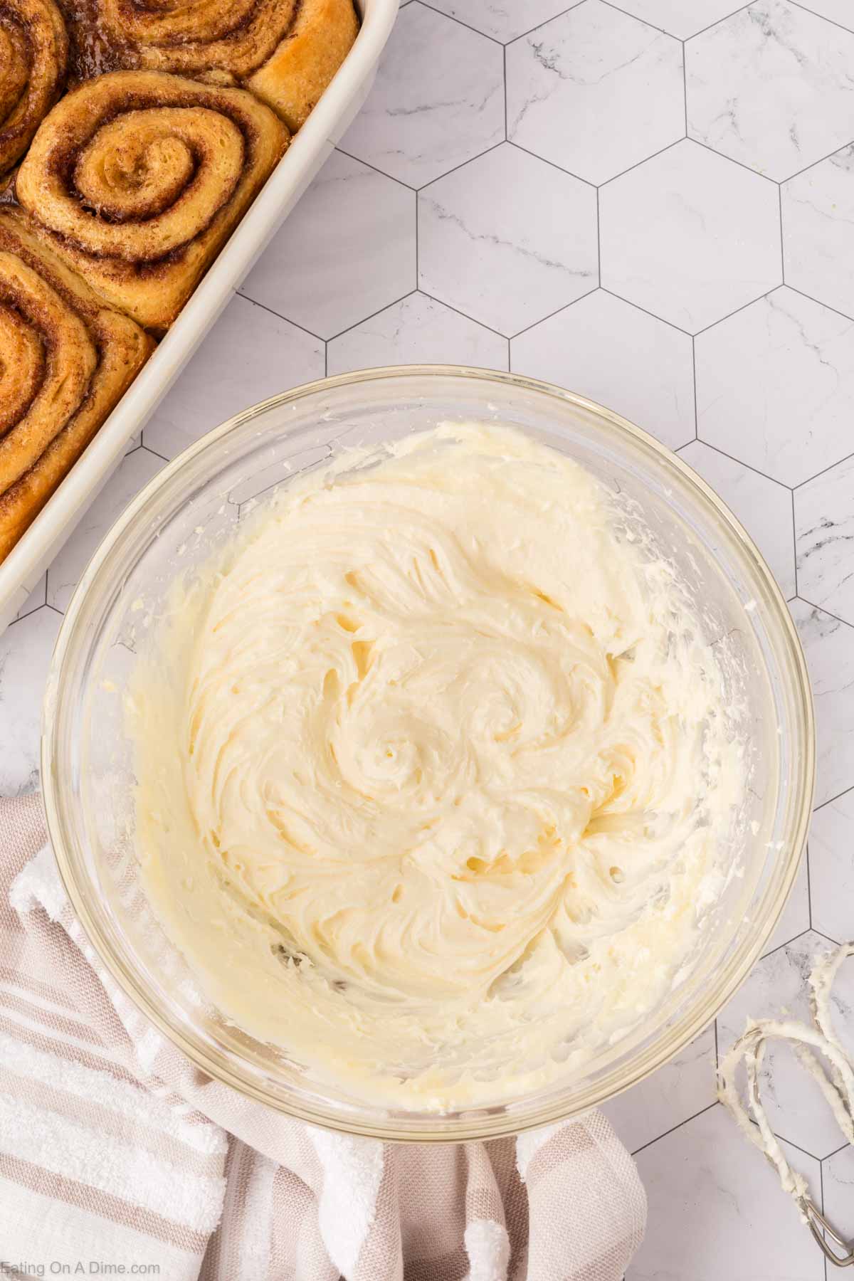 A glass bowl filled with creamy frosting sits on a white hexagon-tiled counter next to a dish of baked cinnamon rolls, inspired by the viral TikTok Cinnamon Rolls Recipe, and a striped kitchen towel.
