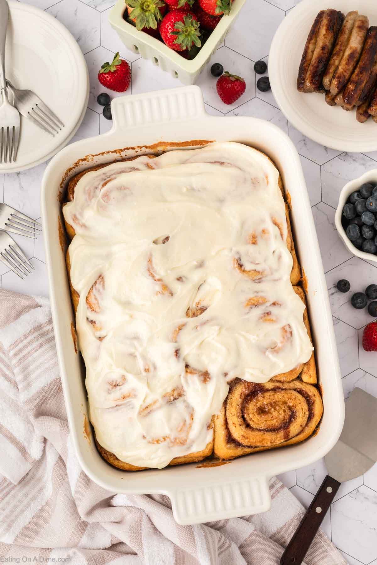 A baking dish filled with TikTok Cinnamon Rolls Recipe topped with creamy white icing, with one roll partially visible without icing. Surrounding the dish are fresh strawberries, blueberries, sausage links, and a striped towel.