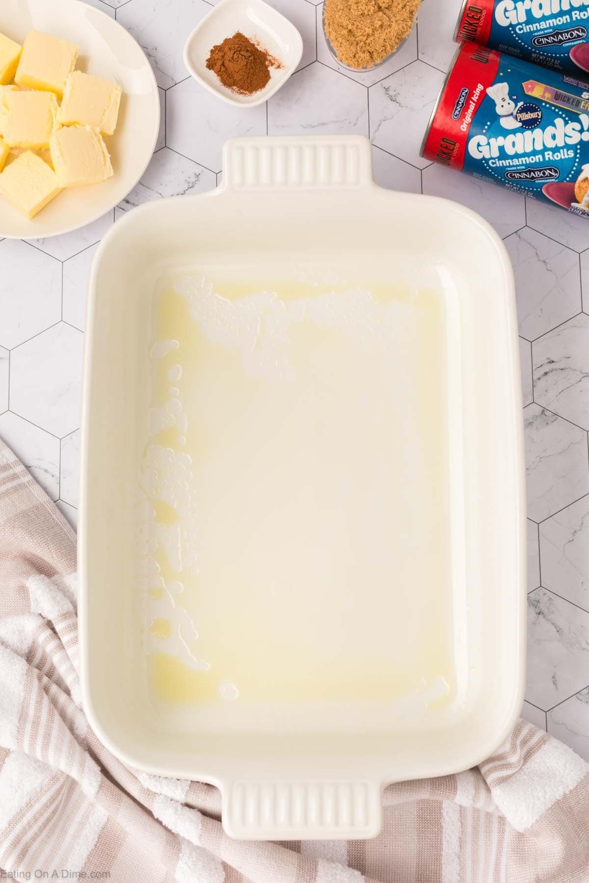 A greased rectangular baking dish sits on a white countertop surrounded by cinnamon, brown sugar, butter, and canned cinnamon rolls—key ingredients for the popular TikTok Cinnamon Rolls Recipe. A striped towel is partially visible in the corner.