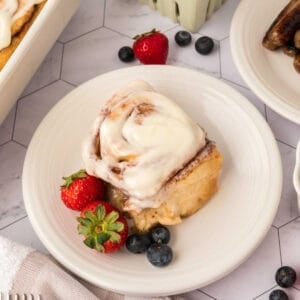 A cinnamon roll made with the TikTok Cinnamon Rolls Recipe is topped with cream cheese frosting on a white plate, garnished with fresh strawberries and blueberries. Some sausages and more fruit are visible in the background.