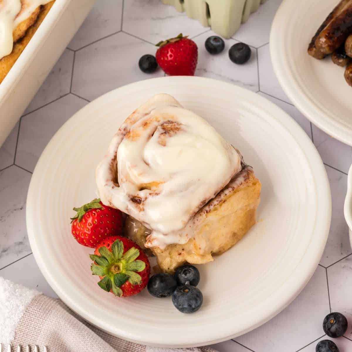 A cinnamon roll made with the TikTok Cinnamon Rolls Recipe is topped with cream cheese frosting on a white plate, garnished with fresh strawberries and blueberries. Some sausages and more fruit are visible in the background.