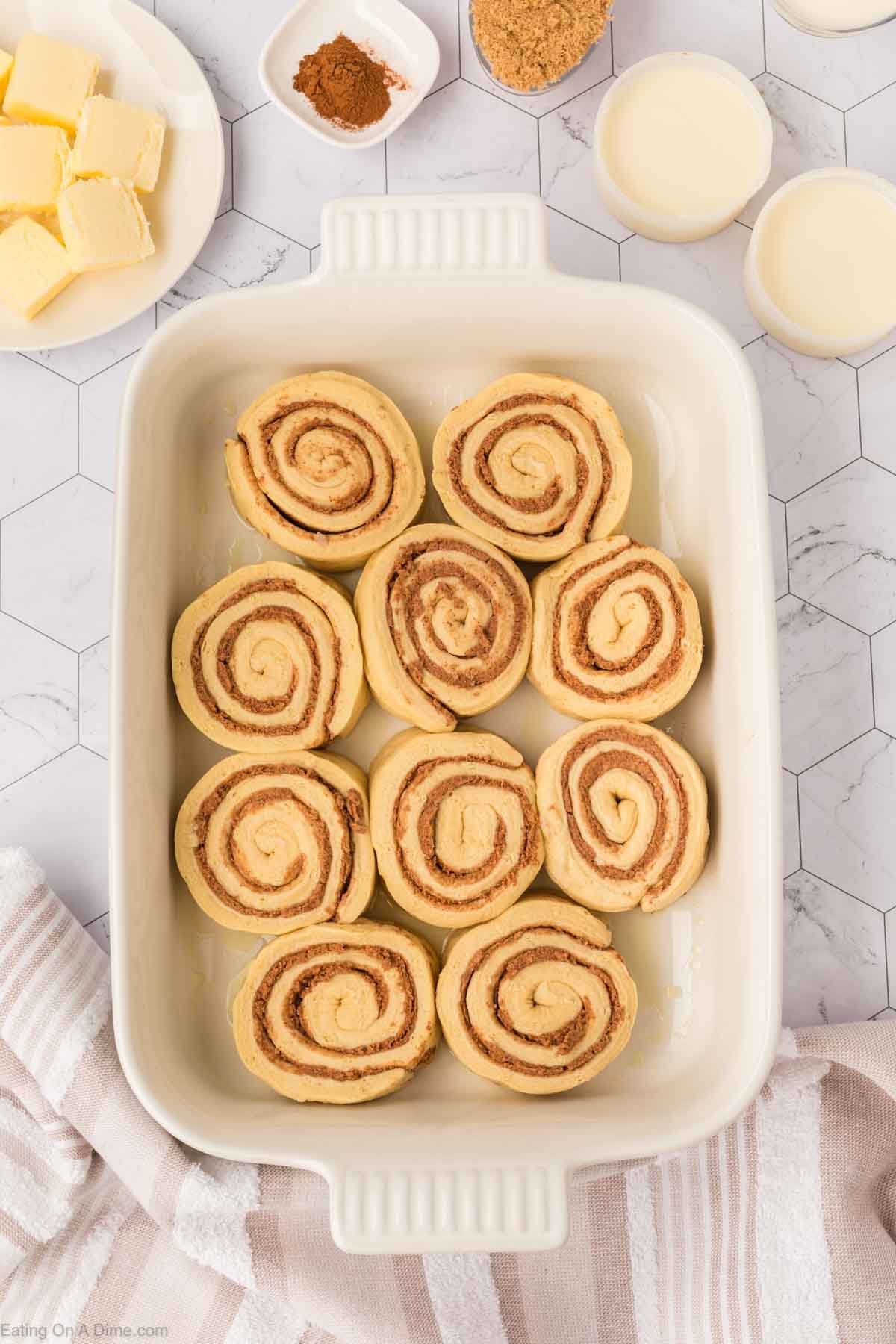 A white baking dish holds nine unbaked TikTok Cinnamon Rolls Recipe favorites. Nearby are small bowls with brown sugar, cinnamon, butter cubes, and two bowls of cream on a marble surface with a striped towel beside the dish.