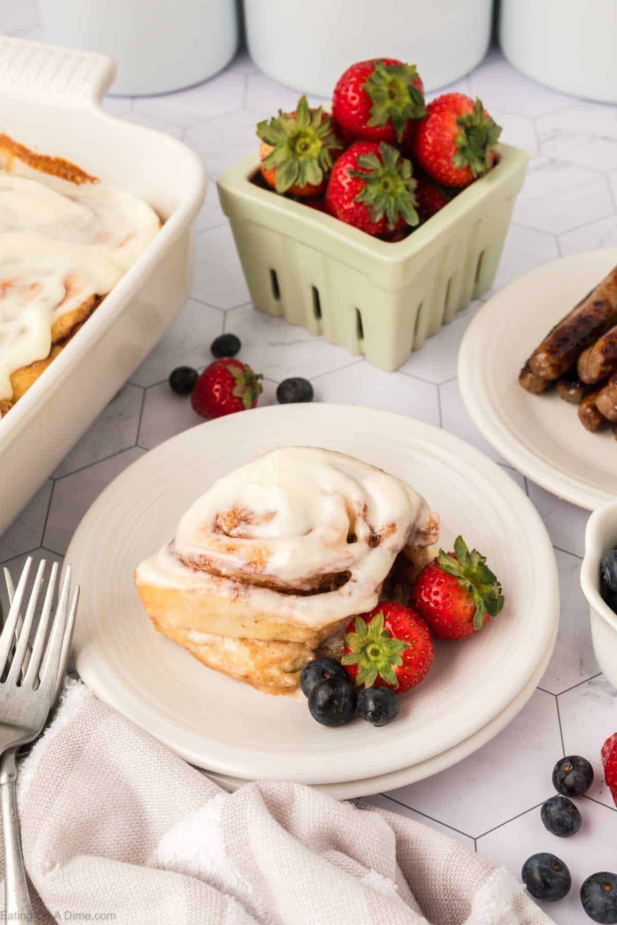 A plate with a cinnamon roll topped with icing, fresh strawberries, and blueberries sits next to a fork. In the background are a basket of strawberries, a dish of TikTok Cinnamon Rolls Recipe, and a plate of breakfast sausages.
