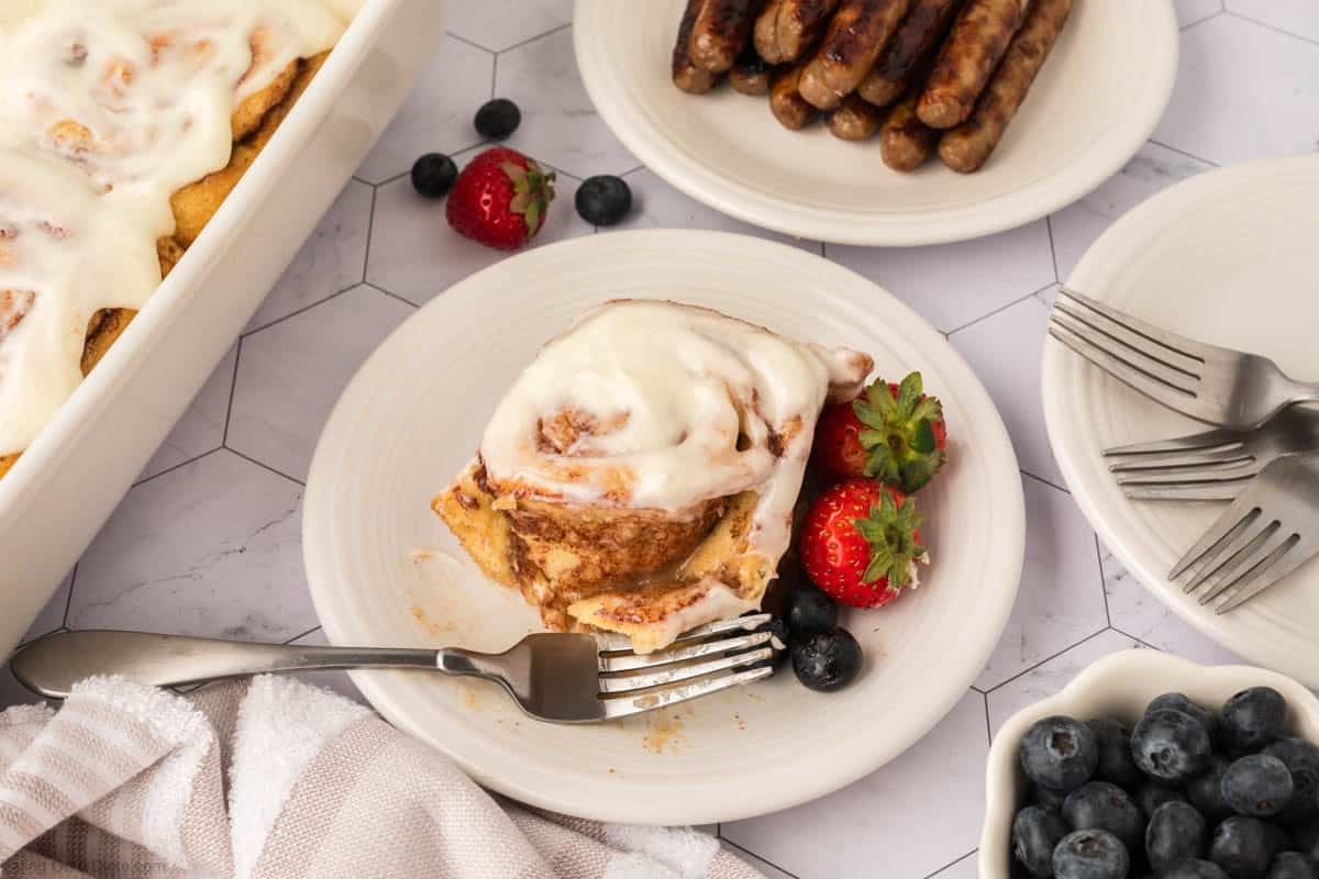 A plate with a frosted cinnamon roll—made using the TikTok Cinnamon Rolls Recipe—strawberries, and blueberries sits next to a fork. Nearby are plates of breakfast sausages and more cinnamon rolls on a light hexagon-patterned table.
