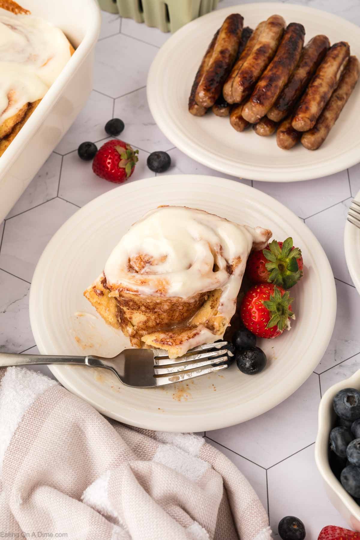 A frosted cinnamon roll, made using the TikTok Cinnamon Rolls Recipe, sits on a plate with strawberries and blueberries, a fork beside it. Nearby are cooked sausages and a partially visible dish towel on the table.