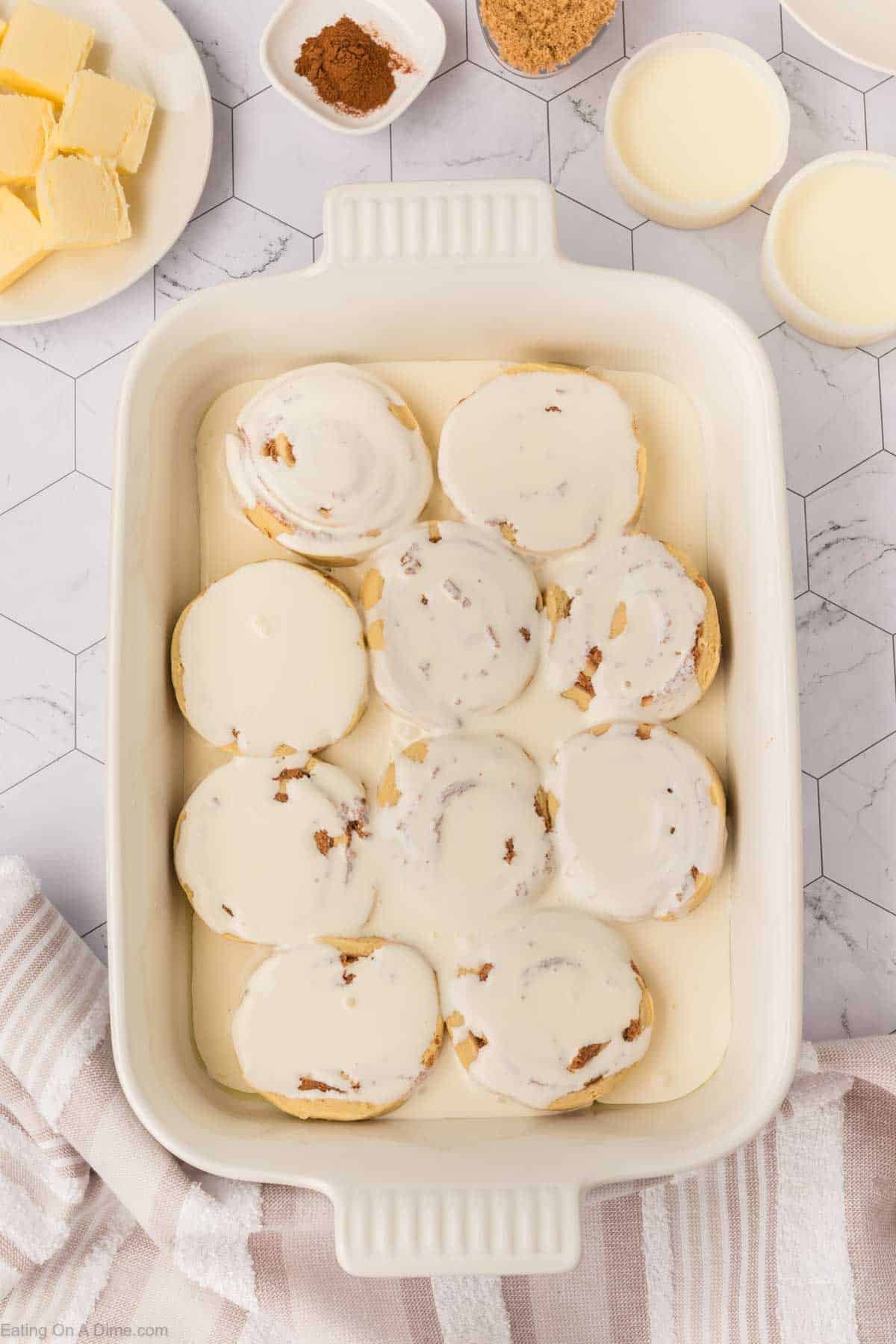 A baking dish filled with nine cinnamon rolls covered in creamy icing, surrounded by bowls of butter, cinnamon, brown sugar, and cream on a white hexagon-tiled surface—perfect for trying the latest TikTok Cinnamon Rolls Recipe.