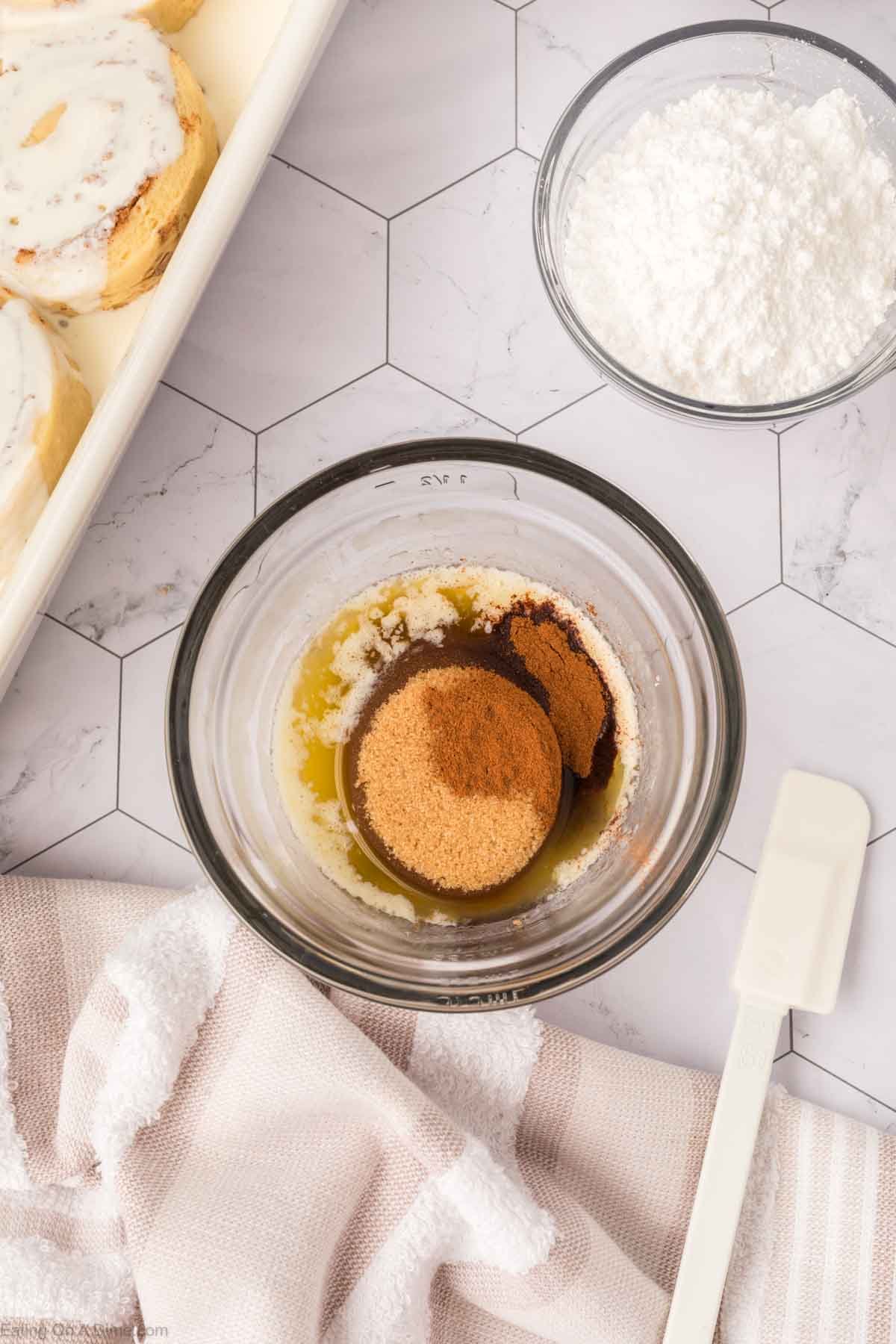 A glass bowl with melted butter, brown sugar, and cinnamon sits on a hexagon-tiled surface next to a white spatula, a bowl of powdered sugar, a striped towel, and a tray of unbaked TikTok Cinnamon Rolls Recipe with icing.
