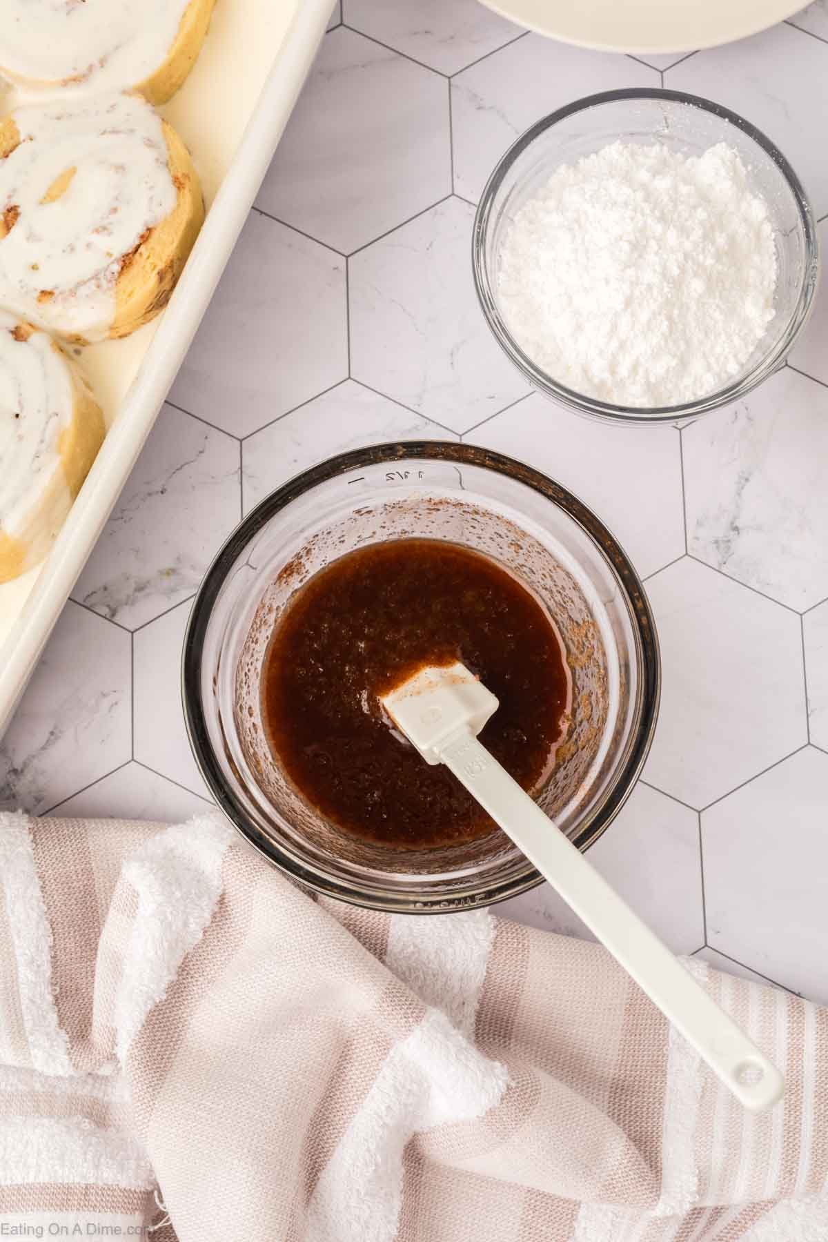 A glass bowl with brown cinnamon glaze and a white spatula sits on a hexagon-tiled surface. Nearby are a bowl of powdered sugar, a tray of iced cinnamon rolls made from the viral TikTok Cinnamon Rolls Recipe, and a beige-striped kitchen towel.