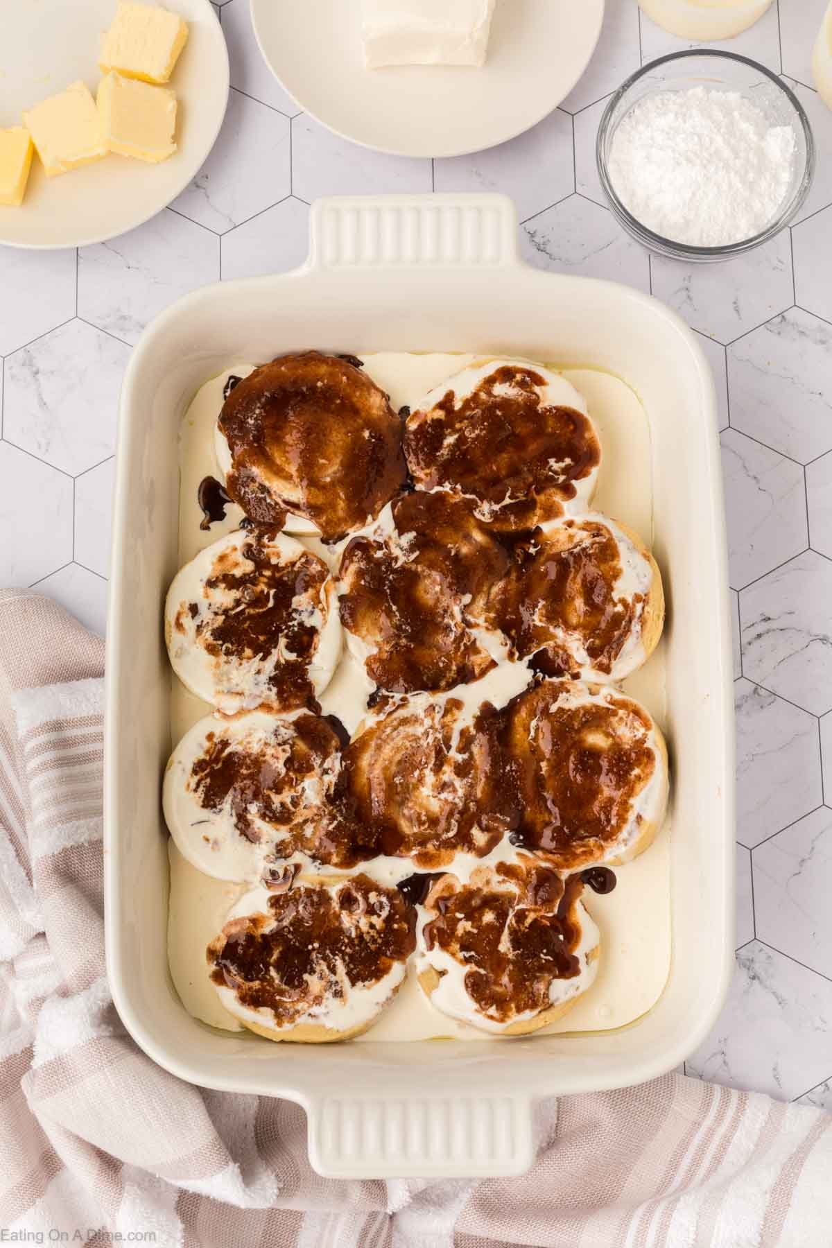 A white baking dish filled with unbaked cinnamon rolls, topped with cinnamon sugar, sits on a marble countertop surrounded by butter, powdered sugar, and cream cheese—perfect for trying out that viral TikTok Cinnamon Rolls Recipe.