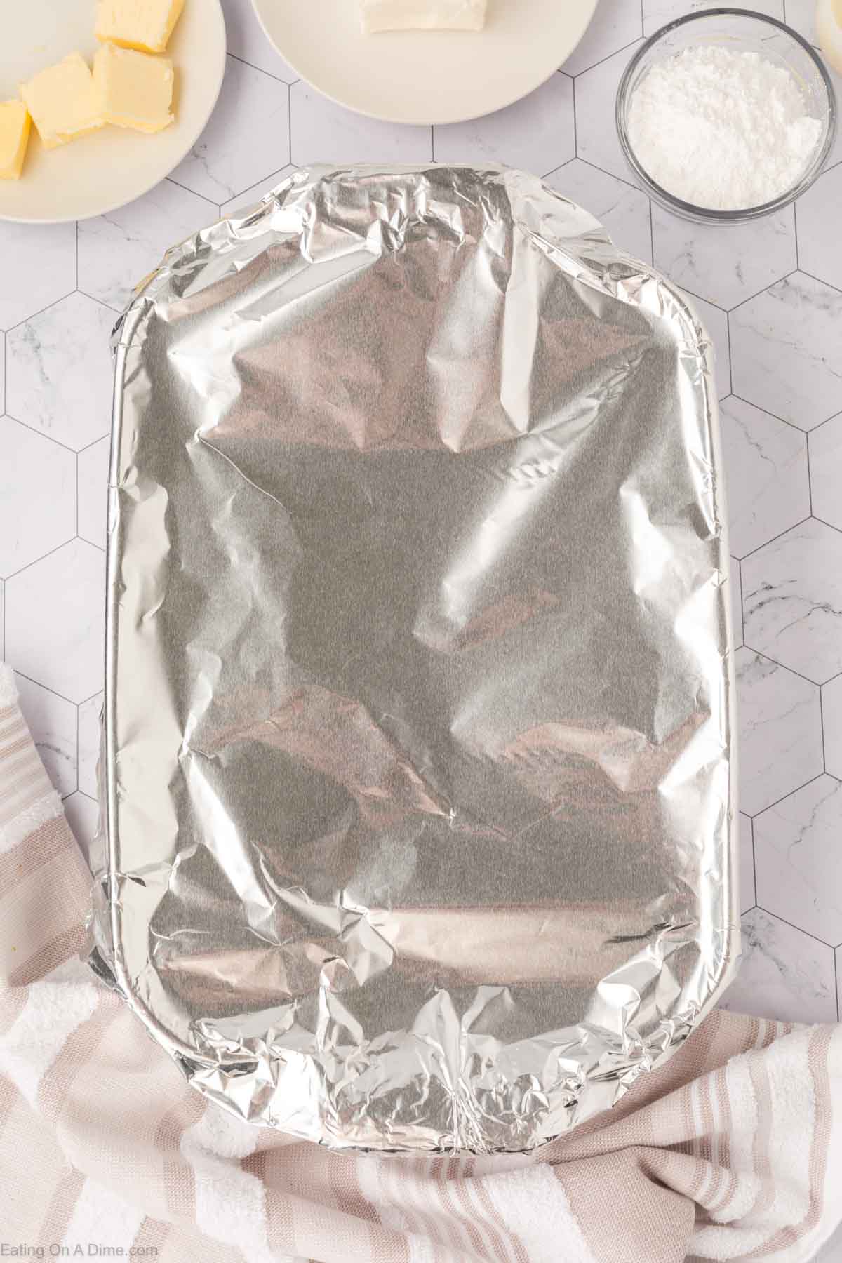 A rectangular baking dish covered with aluminum foil sits on a white hexagon-tiled surface, ready for the TikTok Cinnamon Rolls Recipe. Surrounding it are a plate with butter, a bowl of powdered sugar, and a beige-striped towel.