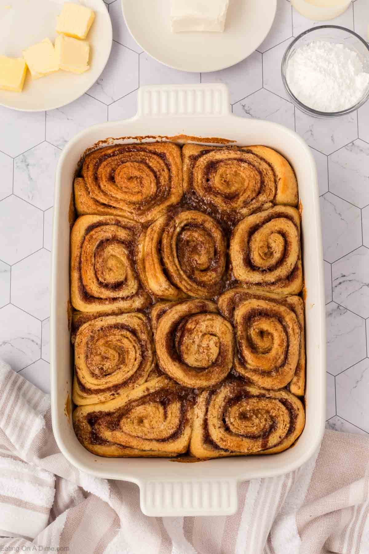 A white baking dish filled with golden-brown cinnamon rolls, made using the viral TikTok Cinnamon Rolls Recipe, sits on a marble countertop, surrounded by small plates of butter, cream cheese, and powdered sugar. A striped towel is nearby.