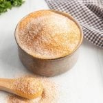 A ceramic bowl filled with a coarse, orange and white seasoning blend—reminiscent of a lawry's seasoned salt recipe—sits on a white surface, with a wooden spoon holding some of the mix. A checkered cloth rests in the background.