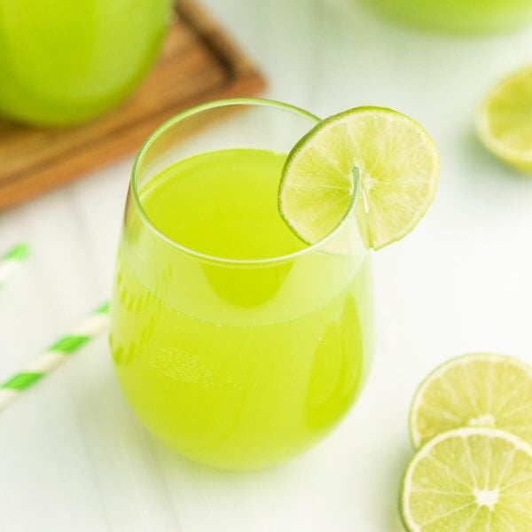 A glass of bright green lime juice garnished with a lime wheel, surrounded by lime halves and a green-striped straw on a white surface