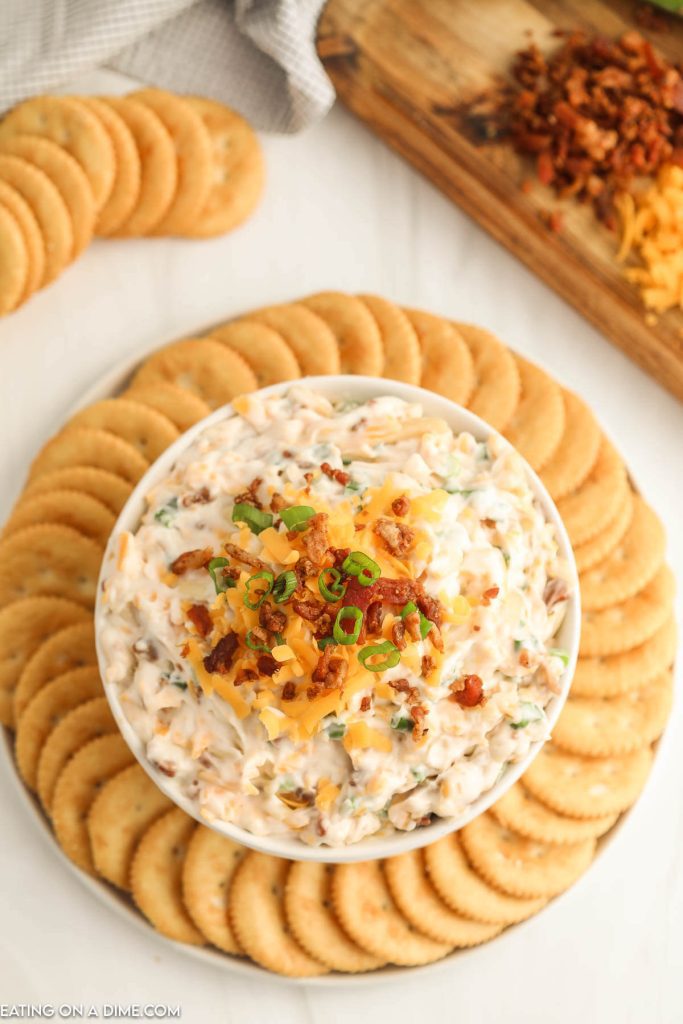 Million dollar dip in a white bowl topped with cheese, bacon, and green onions with a side of crackers