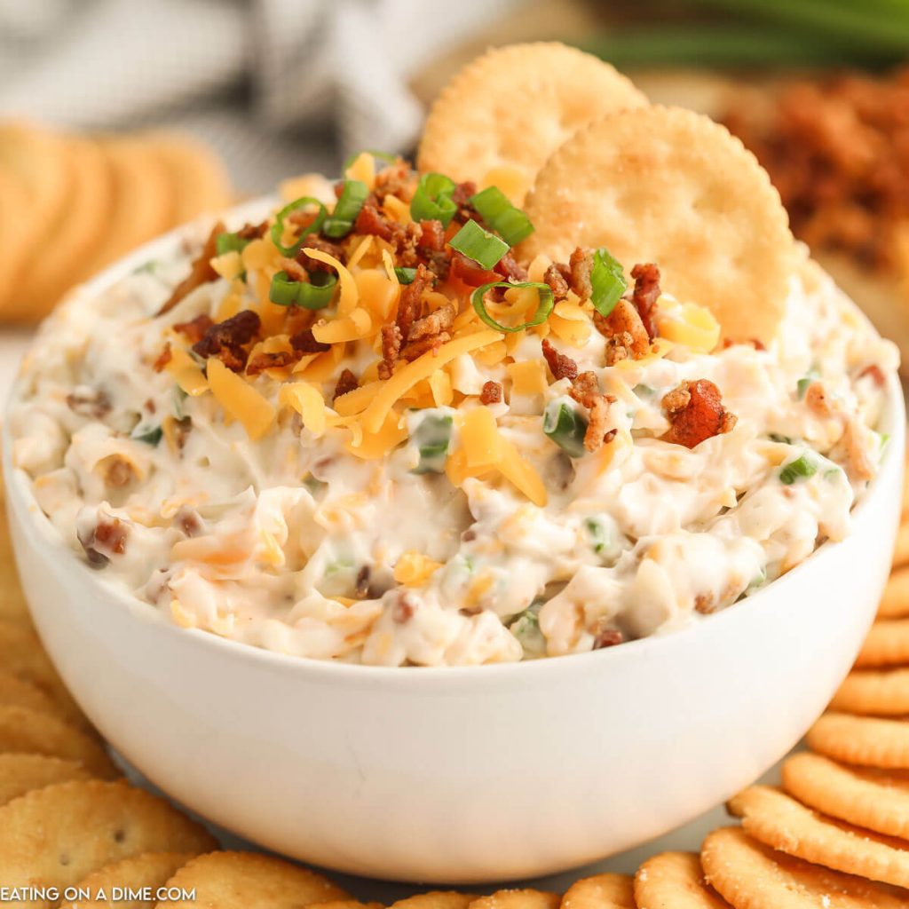 Million dollar dip in a white bowl topped with cheese, bacon, and green onions with a side of crackers