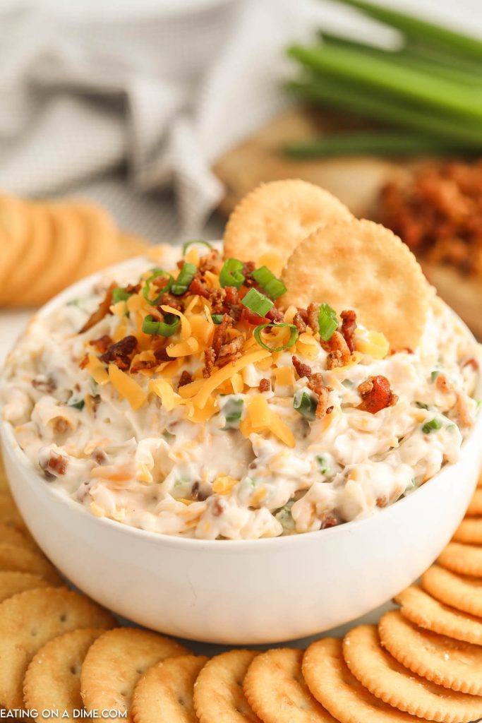 Million dollar dip in a white bowl topped with cheese, bacon, and green onions with a side of crackers
