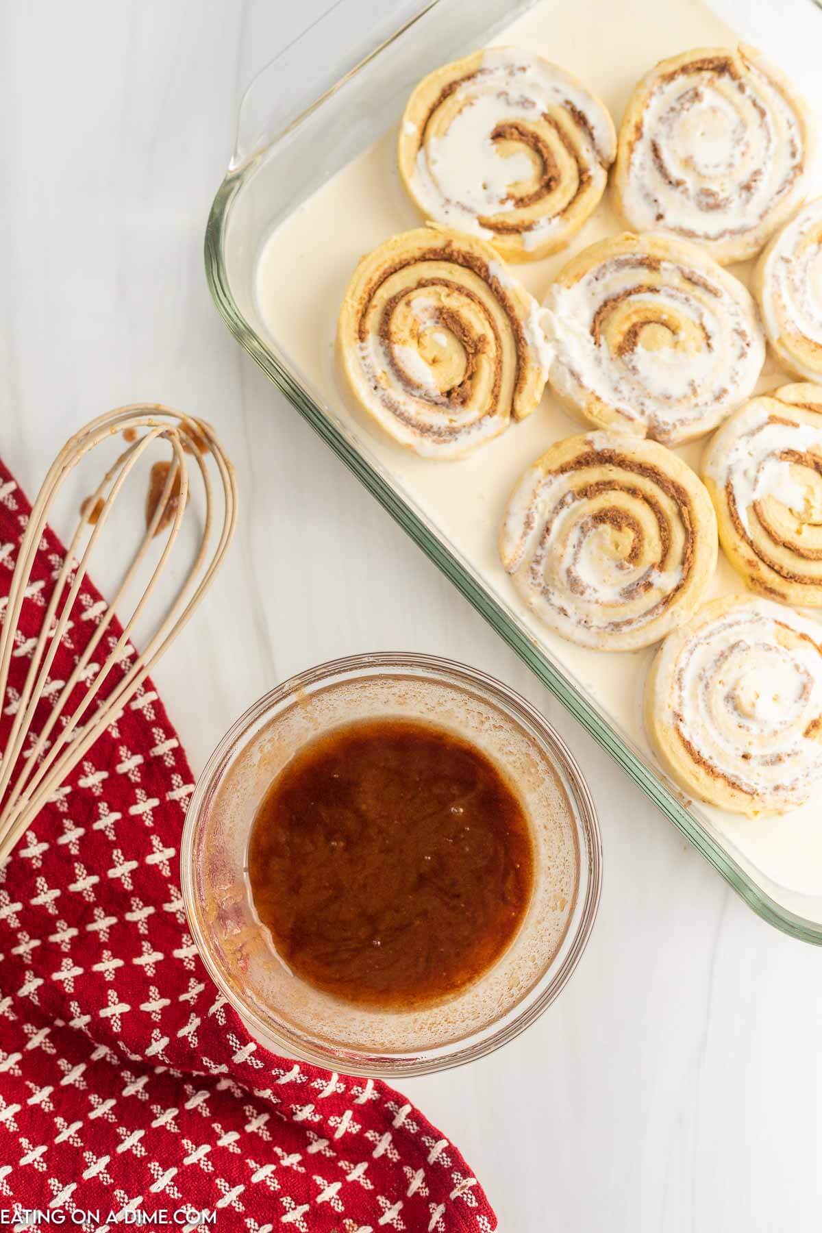 Bowl of cinnamon mixture on the side of a glass baking dish with uncooked cinnamon rolls covered with heavy cream