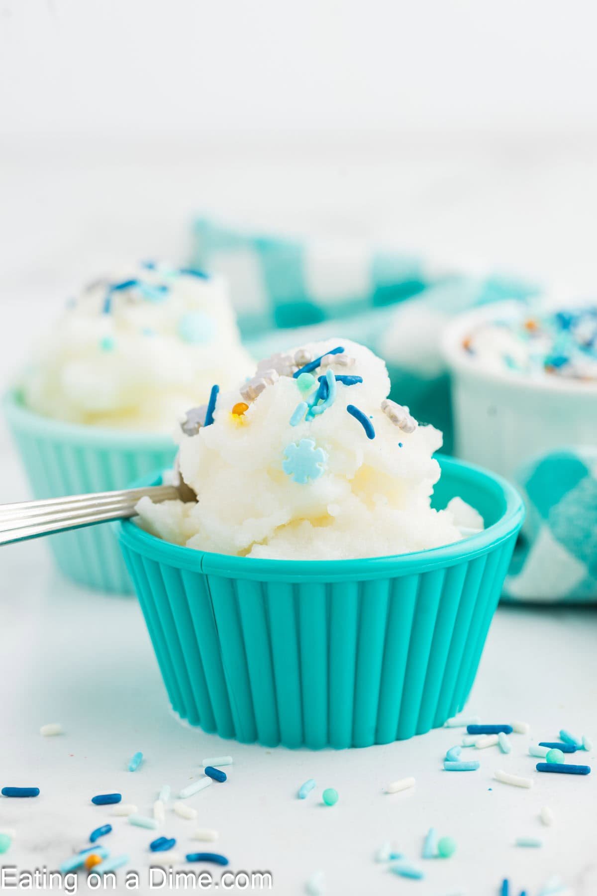 A teal silicone cup filled with white snow ice cream recipe, topped with blue and white sprinkles, sits on a white surface. A fork rests inside the cup. More cups and sprinkles are blurred in the background.