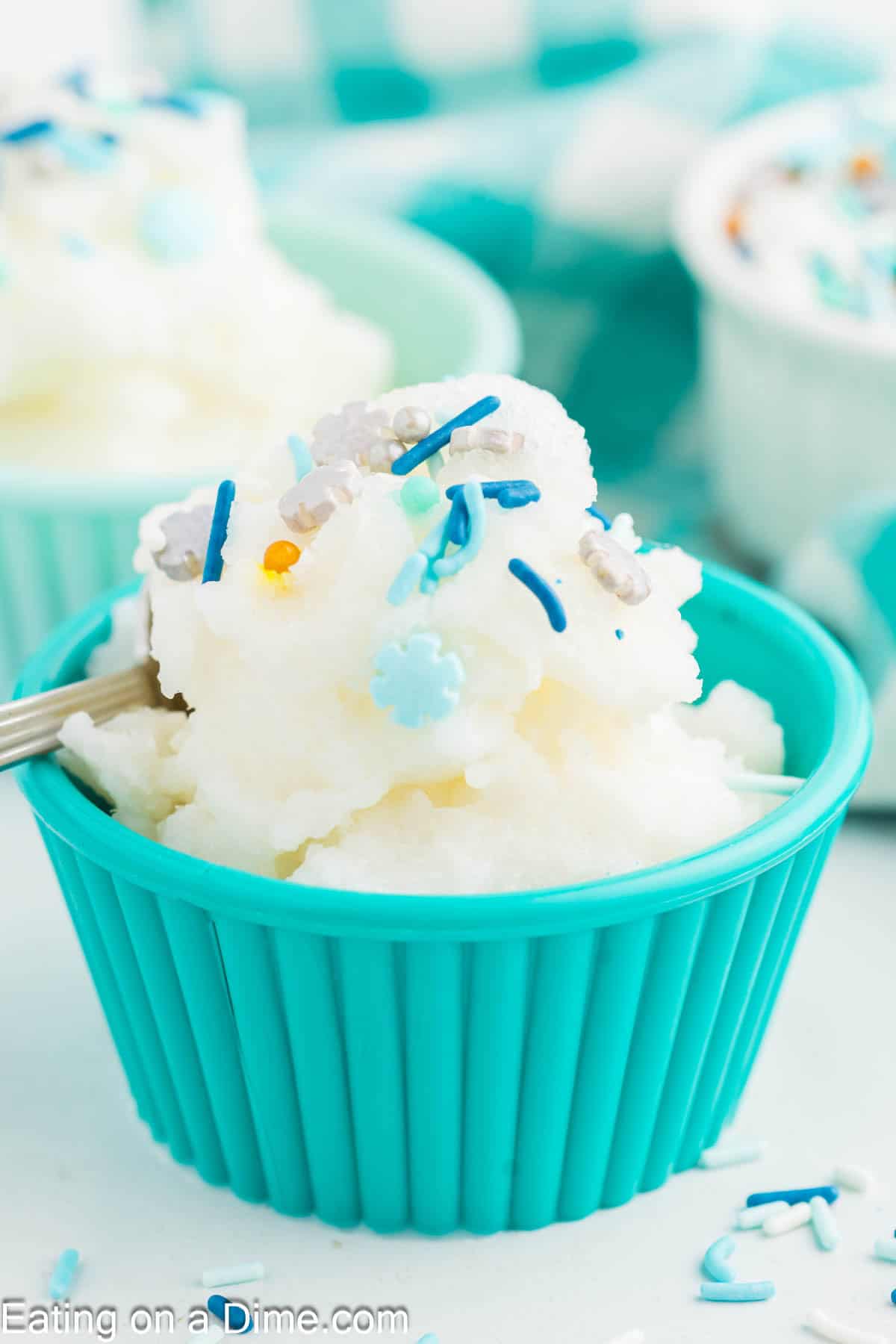 A scoop of white snow ice cream made from a classic snow ice cream recipe sits in a turquoise bowl, topped with blue and silver sprinkles, with a spoon on the side. Another bowl of ice cream is blurred in the background.
