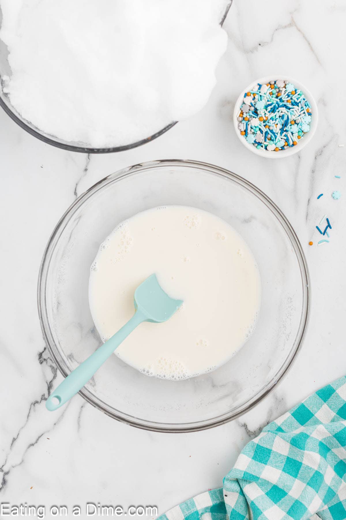 A glass bowl filled with a creamy white snow ice cream recipe and a blue spatula sits on a marble surface, next to a bowl of white fluff, a small bowl of blue and gold sprinkles, and a teal checkered cloth.