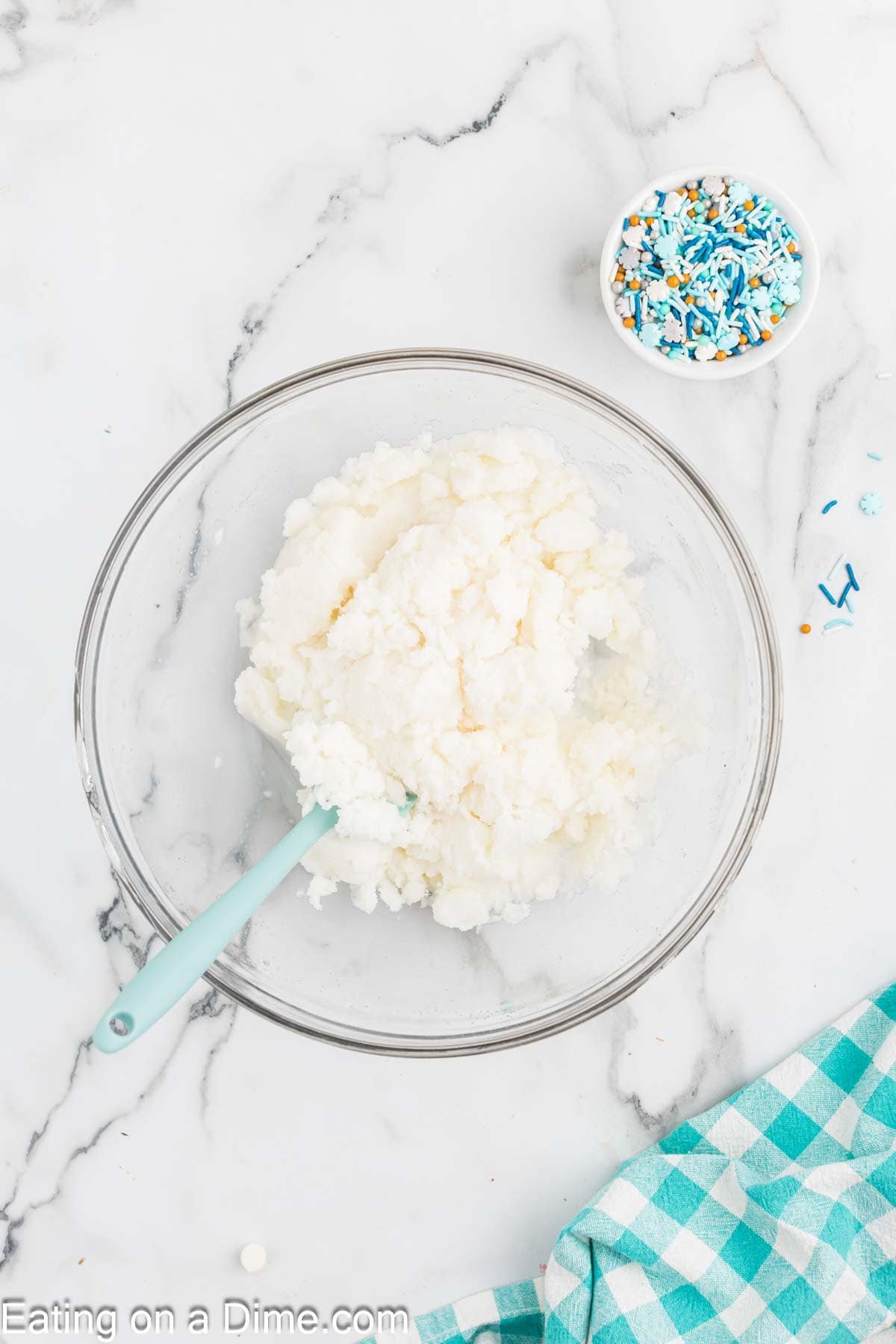 A glass bowl of white frosting or icing, perfect for a snow ice cream recipe, sits on a marble surface with a blue spatula. Nearby are blue, white, and gold sprinkles in a small bowl and a teal checkered cloth partially visible.