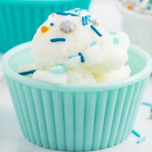 A scoop of white snow ice cream recipe topped with blue and silver sprinkles is served in a light blue, ribbed bowl. Some sprinkles are scattered around the bowl on a white surface.