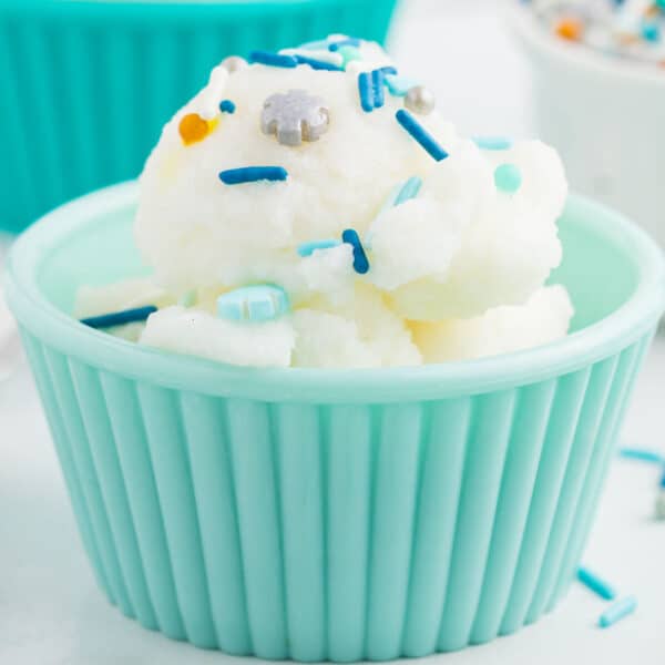 A scoop of white snow ice cream recipe topped with blue and silver sprinkles is served in a light blue, ribbed bowl. Some sprinkles are scattered around the bowl on a white surface.