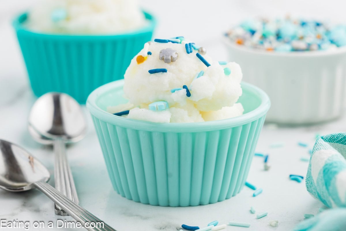A small blue bowl holds a serving of white snow ice cream, topped with blue and white sprinkles. Nearby are metal spoons, a napkin, and another bowl filled with more sprinkles—perfect for trying out your favorite snow ice cream recipe.