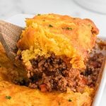 A wooden spoon lifts a serving of Sloppy Joe Cornbread Casserole with a golden, cheesy cornbread topping and savory ground meat filling from a baking dish.