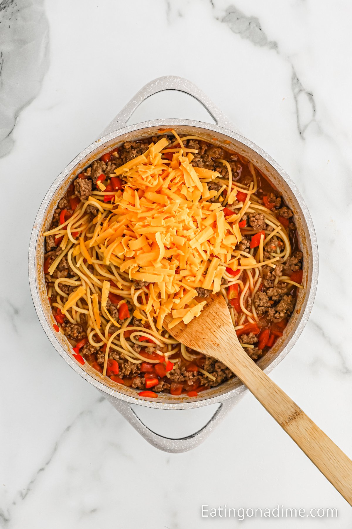 Topping the ground beef spaghetting mixture with shredded cheese in a large skillet with a wooden spoon