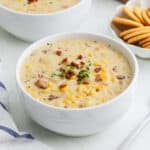 A white bowl filled with creamy corn chowder recipe, topped with bacon bits and herbs, sits on a light surface next to a plate of round crackers. Another bowl of chowder is visible in the background.