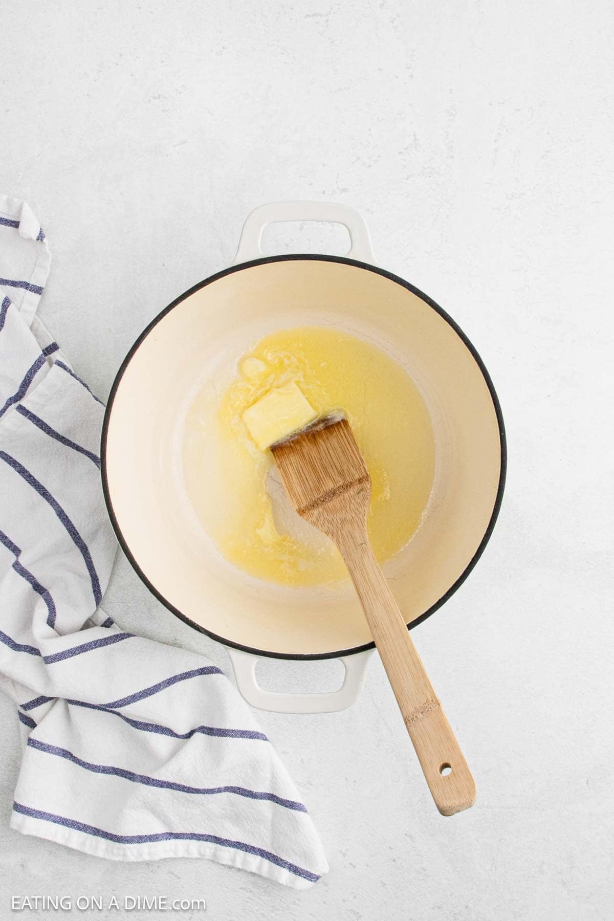 A block of butter melting in a cream-colored pot, stirred with a wooden spatula—the first step in a cozy corn chowder recipe. A white and blue striped kitchen towel sits to the left on a light-colored surface.