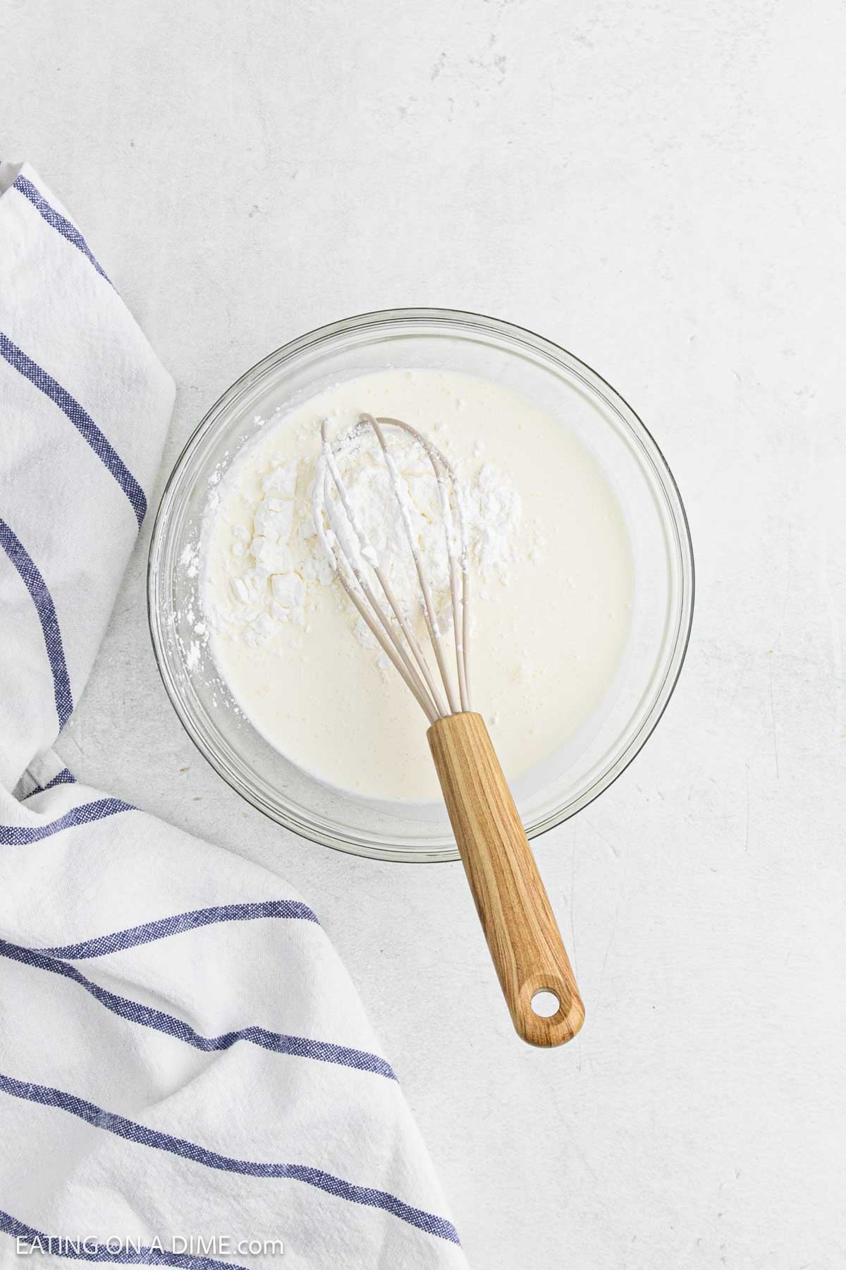 A glass bowl filled with a white batter and a whisk, placed on a light surface next to a white towel with blue stripes—perfect for starting your favorite corn chowder recipe.