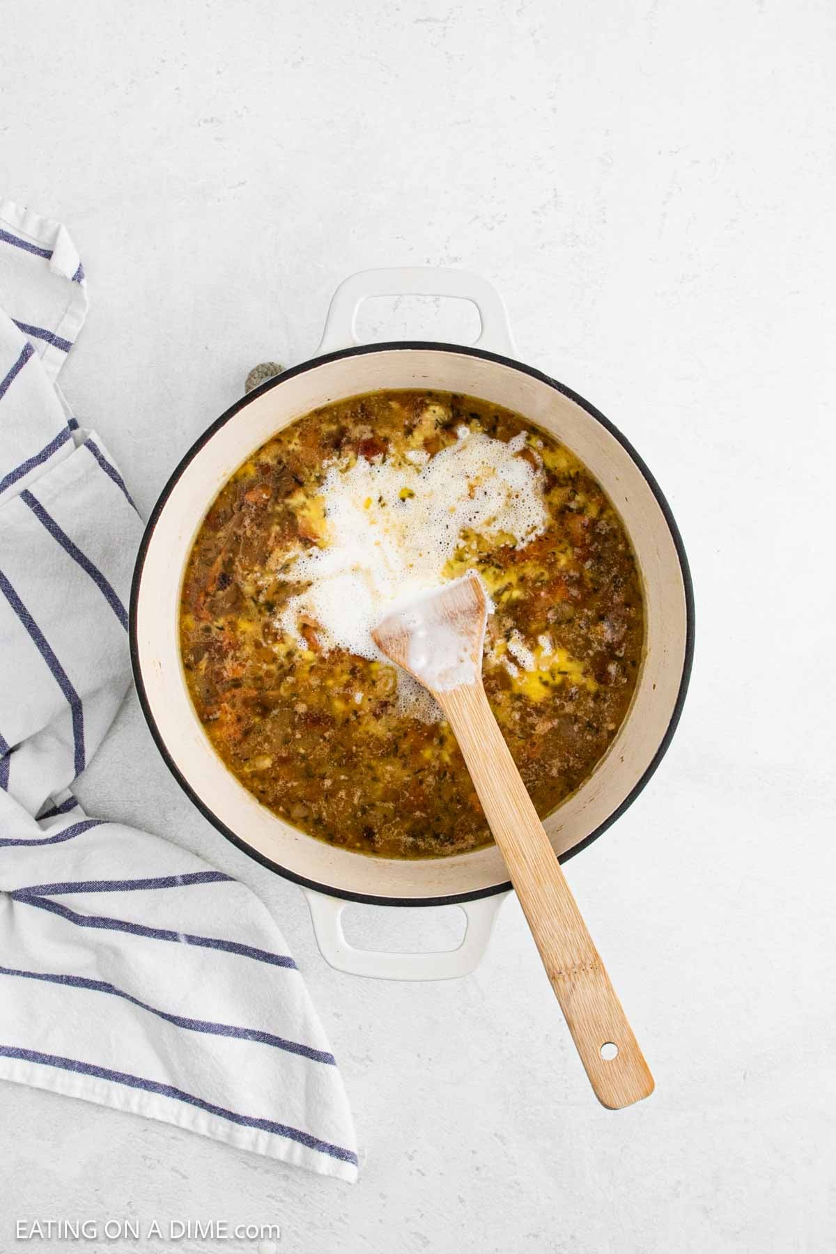 A white pot filled with a seasoned ground meat mixture and a pile of white granulated topping, reminiscent of prepping a corn chowder recipe, is being stirred with a wooden spatula next to a striped kitchen towel on a light surface.