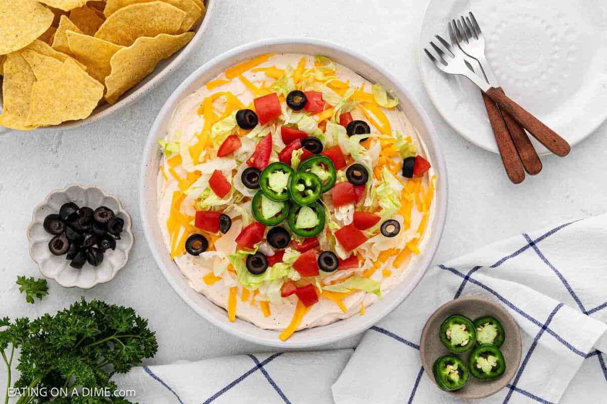 Taco Dip in a white platter topped with shredded lettuce, dice tomatoes, slice jalapenos and black olives with a bowl of tortilla chips on the side