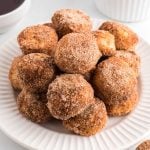 A plate stacked with golden-brown Air Fryer Donut Holes coated in cinnamon sugar on a white background, with a bowl of chocolate sauce partially visible in the corner.