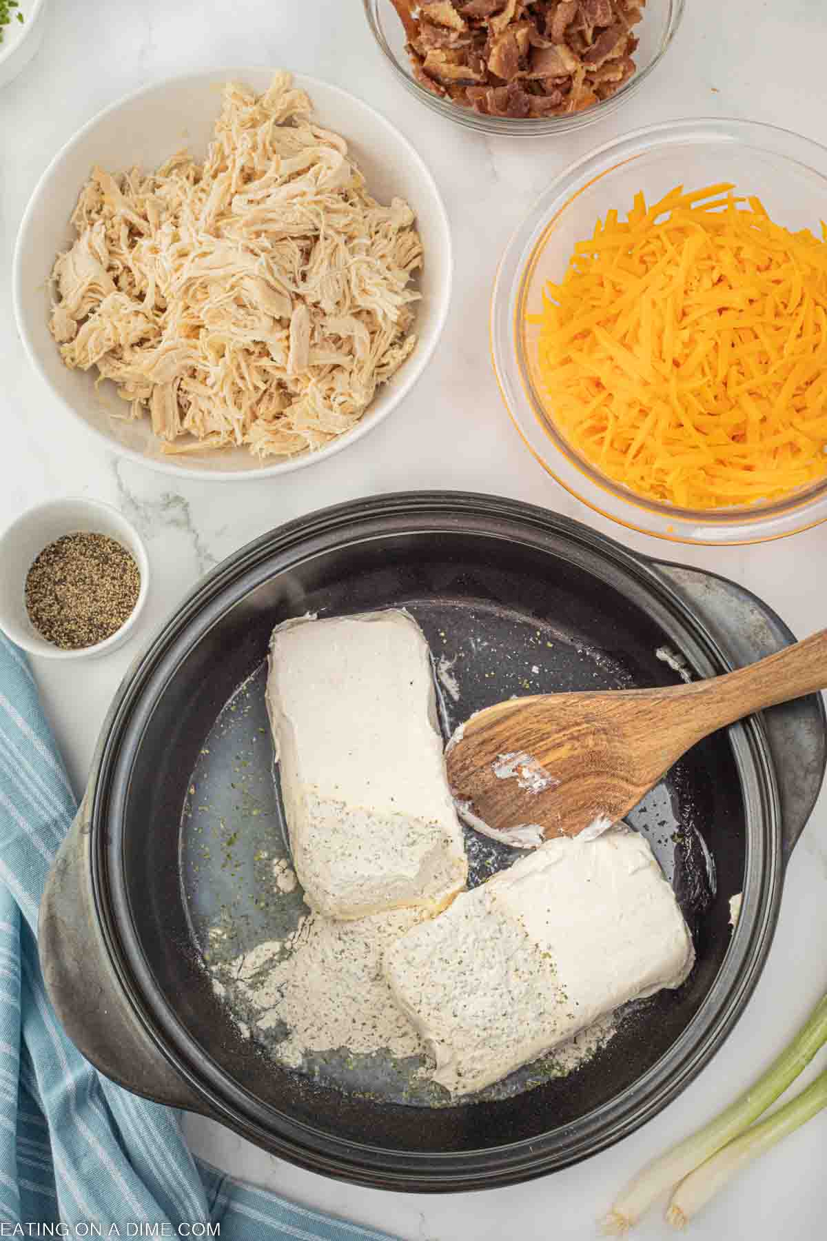 Two cream cheese in a large pot with a wooden spoon with bowl on the side of shredded chicken, shredded cheese, bacon and pepper