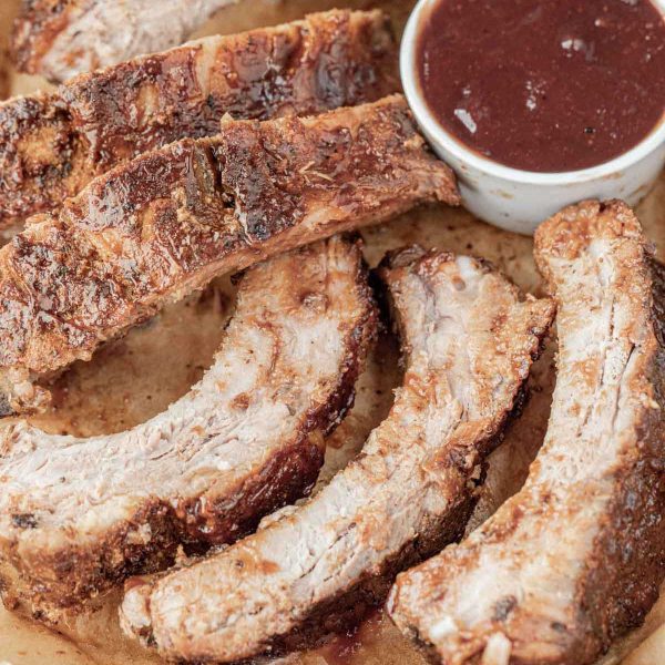 Several pieces of cooked, seasoned Slow Cooker Baby Back Ribs are arranged on parchment paper next to a small cup of barbecue sauce. The ribs have a crispy, browned exterior and are served without any visible garnish.
