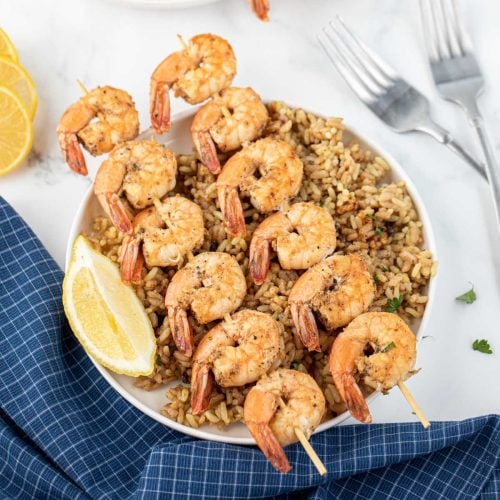 A plate of Texas Roadhouse Grilled Shrimp skewers served over brown rice, garnished with two lemon wedges. Two forks and a blue checkered napkin are next to the plate on a white surface.