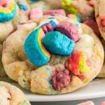 A close-up of a colorful Lucky Charms Cookies sugar cookie topped with vibrant, rainbow-colored marshmallows. The cookie appears soft and thick, with various shades of blue, pink, yellow, and purple marshmallow pieces.
