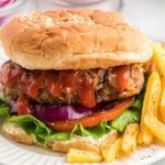 A close-up of a meatloaf burger with lettuce, tomato, red onion, and ketchup in a sesame seed bun, served with a side of golden French fries on a white plate.