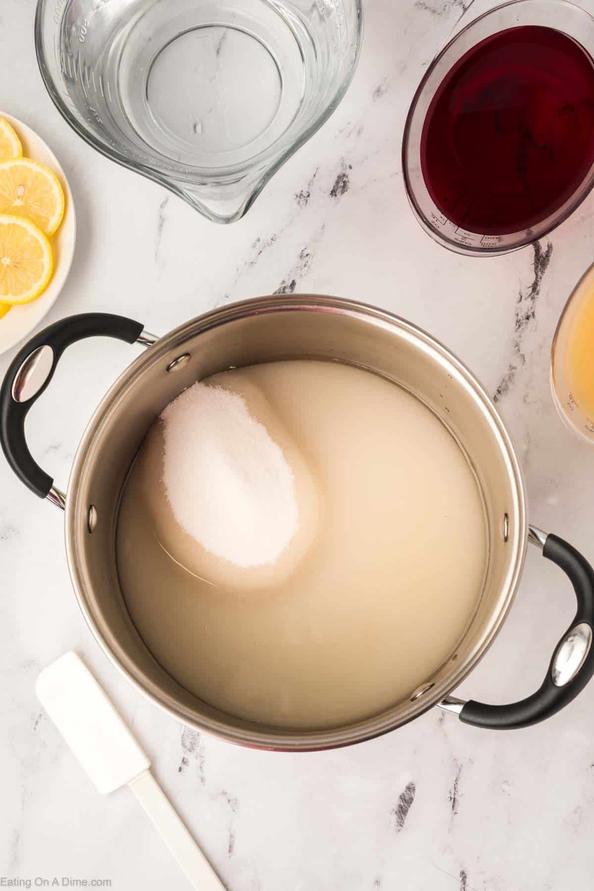 A stainless steel pot on a marble surface contains water and a mound of sugarโperfect beginnings for a Pink Lemonade Recipe. Nearby are a glass measuring cup of water, lemon slices, red and orange liquids in glasses, and a white spatula.