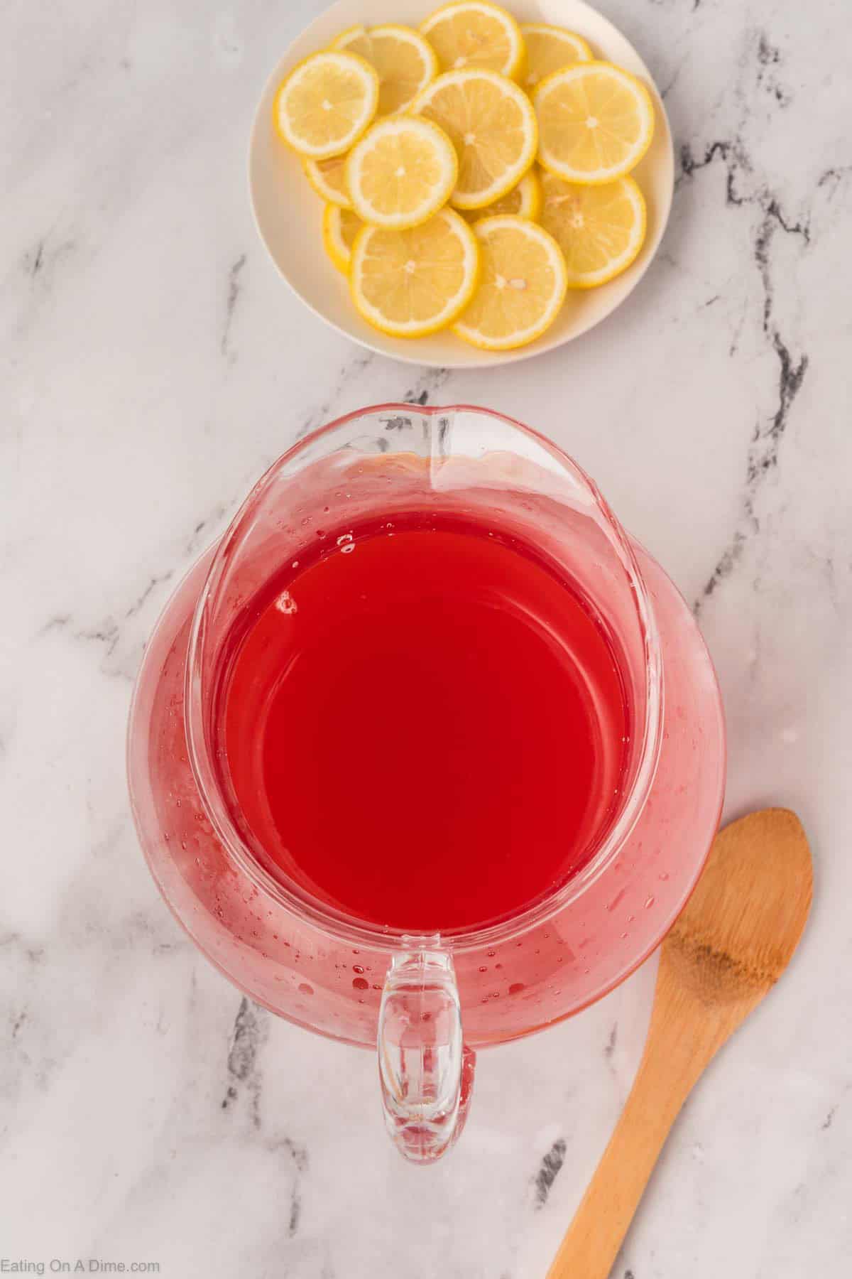A clear pitcher filled with bright red liquid sits on a marble surface next to a wooden spoon and a white plate with sliced lemonsโperfect for showcasing your favorite pink lemonade recipe.