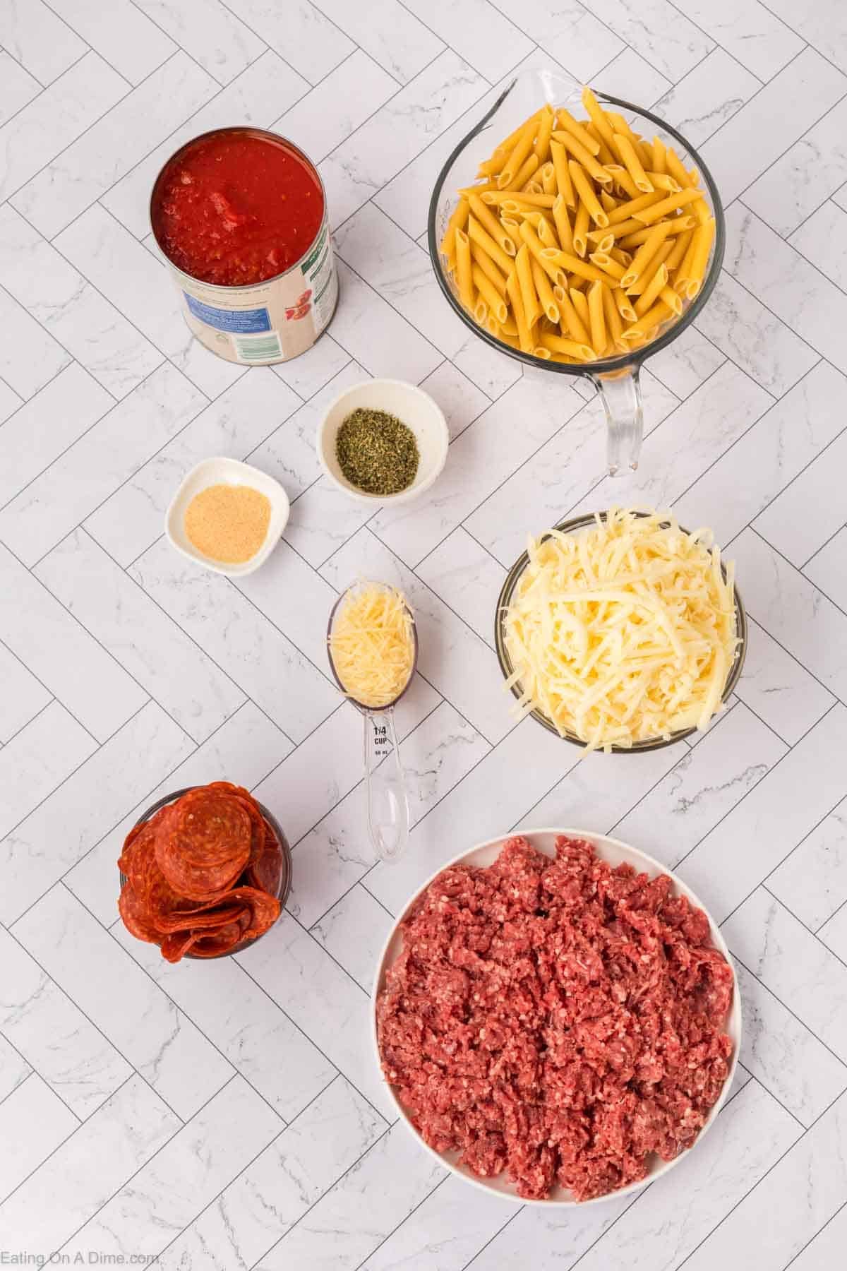 Ingredients for a Pizza Casserole arranged on a white tiled surface: uncooked penne pasta, shredded mozzarella cheese, sliced pepperoni, ground beef, canned tomato sauce, grated parmesan, and dried Italian herbs.