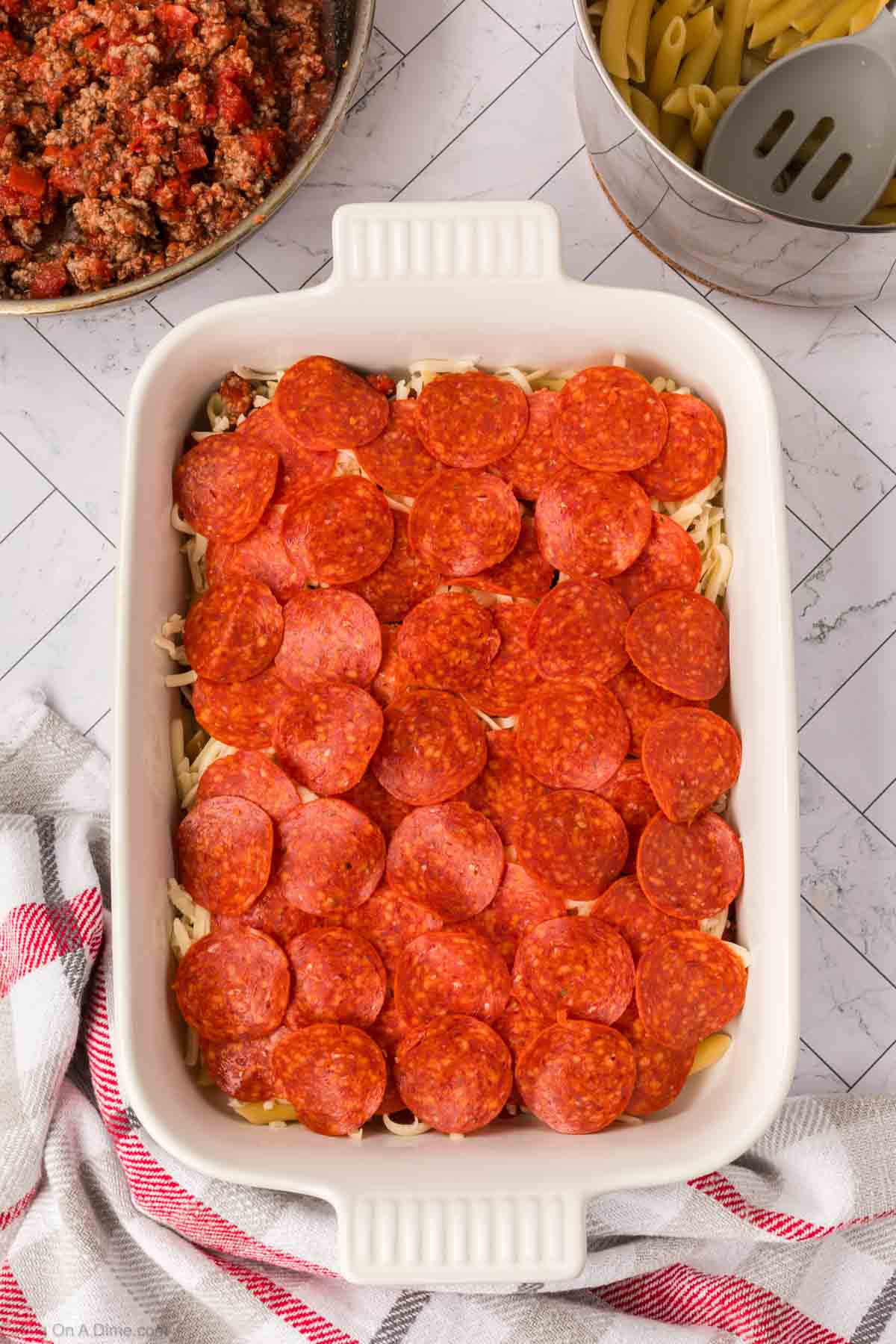 A white baking dish filled with uncooked Pizza Casserole topped with pepperoni slices. Nearby are a bowl of cooked meat sauce, a pot of uncooked pasta, and a red-and-white kitchen towel.