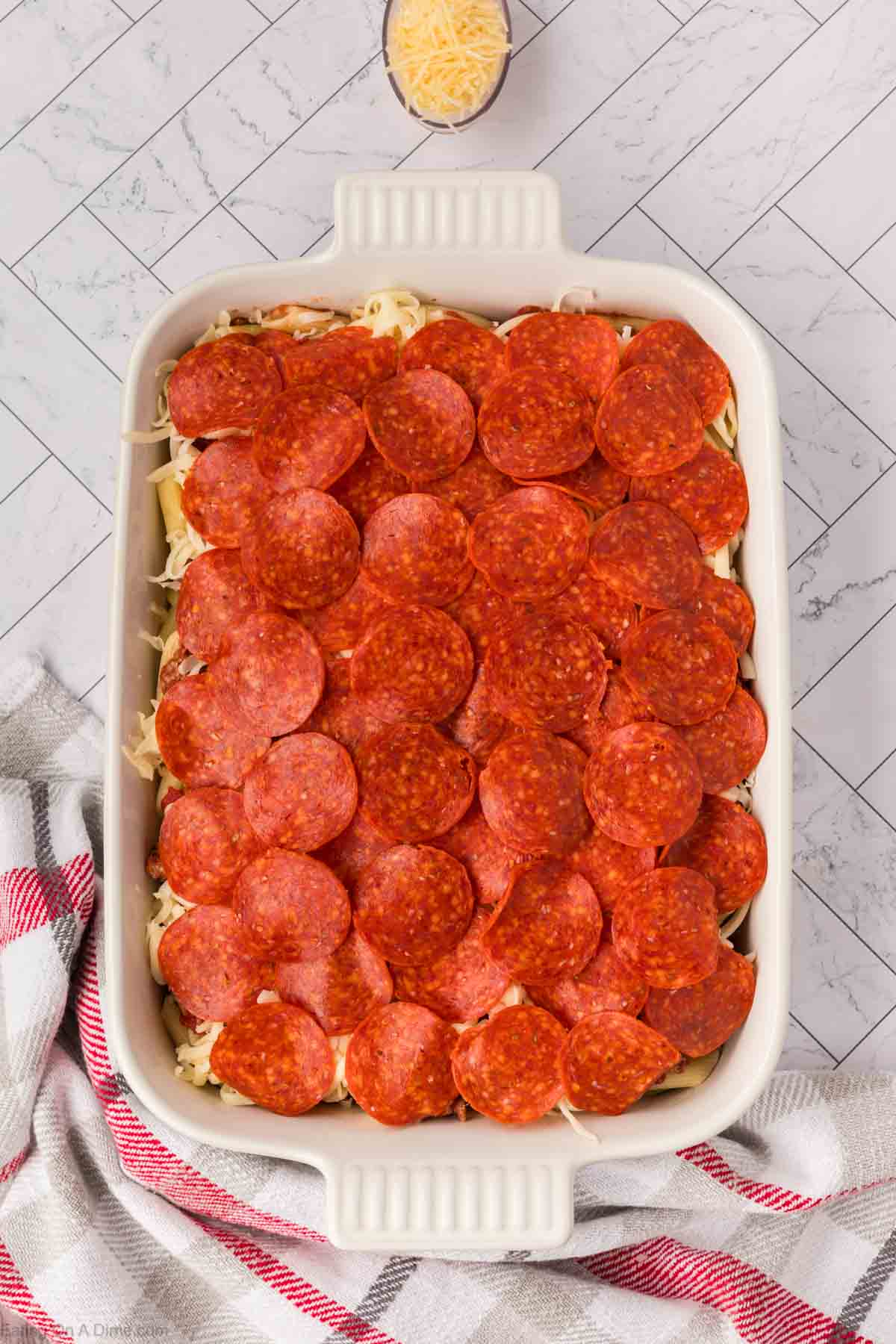 A rectangular white baking dish filled with Pizza Casserole, topped with overlapping slices of pepperoni. A bowl of shredded cheese and a striped towel are nearby on a light, tiled surface.