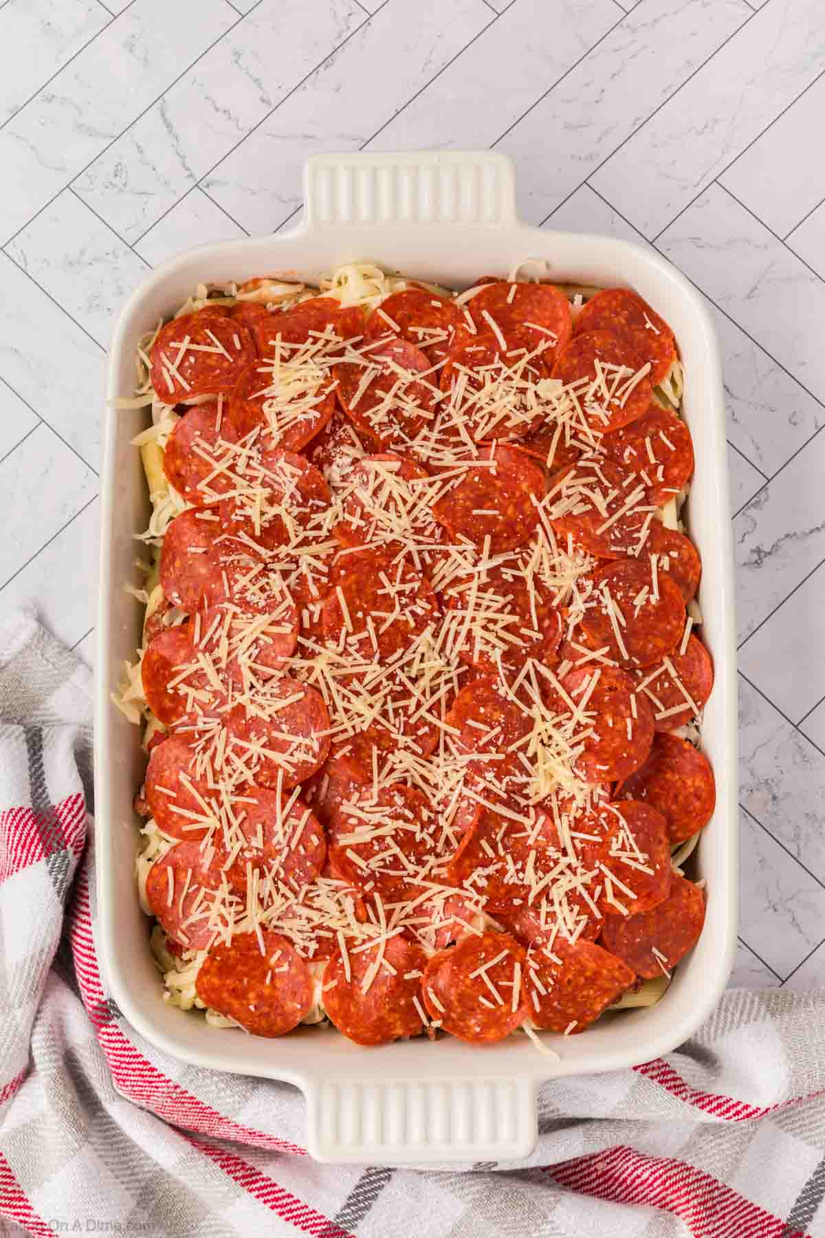 A white baking dish filled with a layered pizza casserole, topped with rows of pepperoni slices and melted cheese, rests on a marble countertop next to a red and white striped kitchen towel.